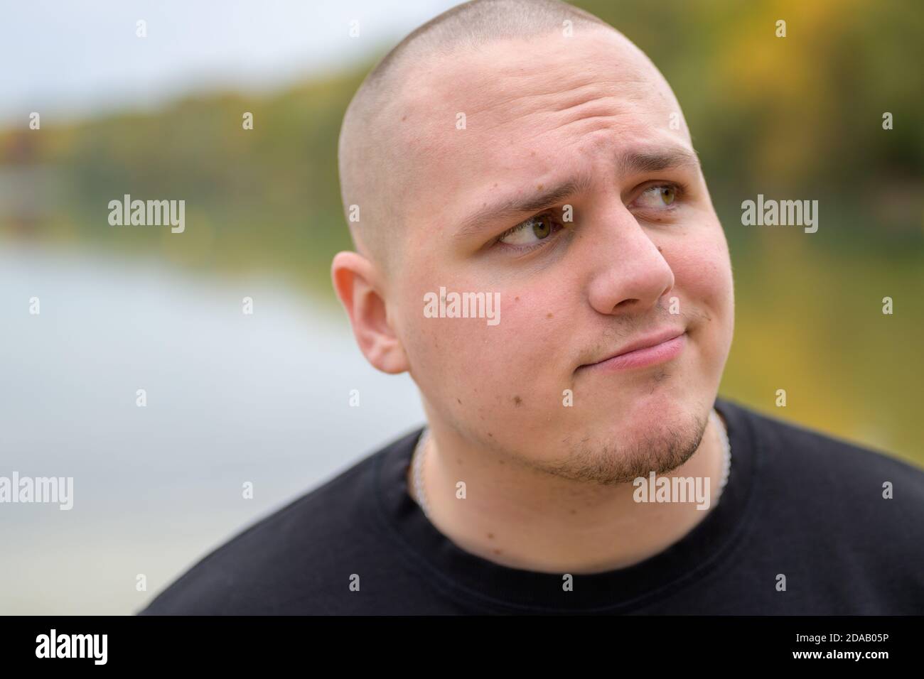 Dubious or confused young man frowning and pursing his lips as he looks up to the side outdoors at an autumn lake in a park Stock Photo