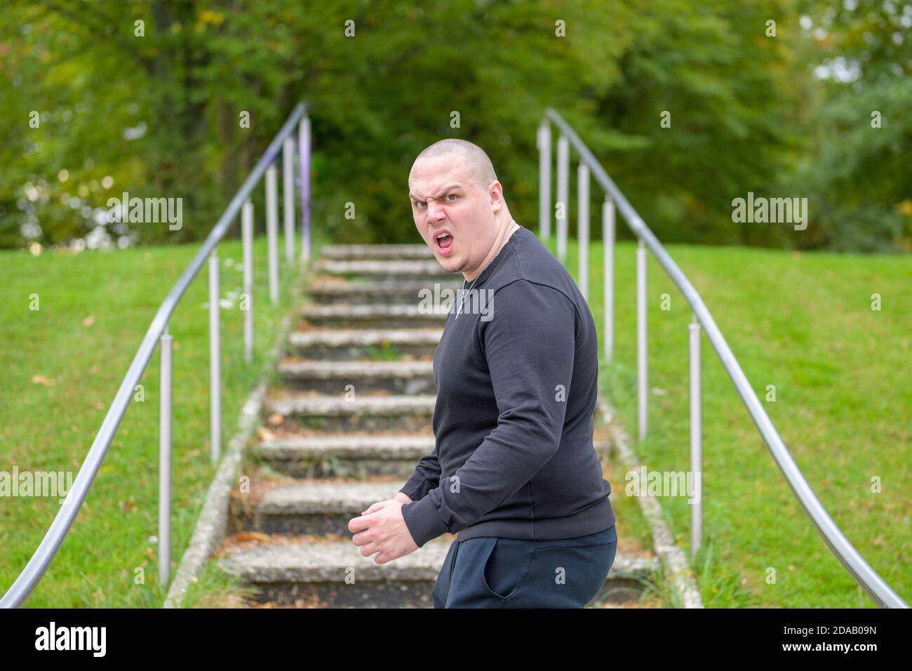 Angry aggressive young man turning to yell at the camera with a look of fury as he walks up steps in a green park Stock Photo