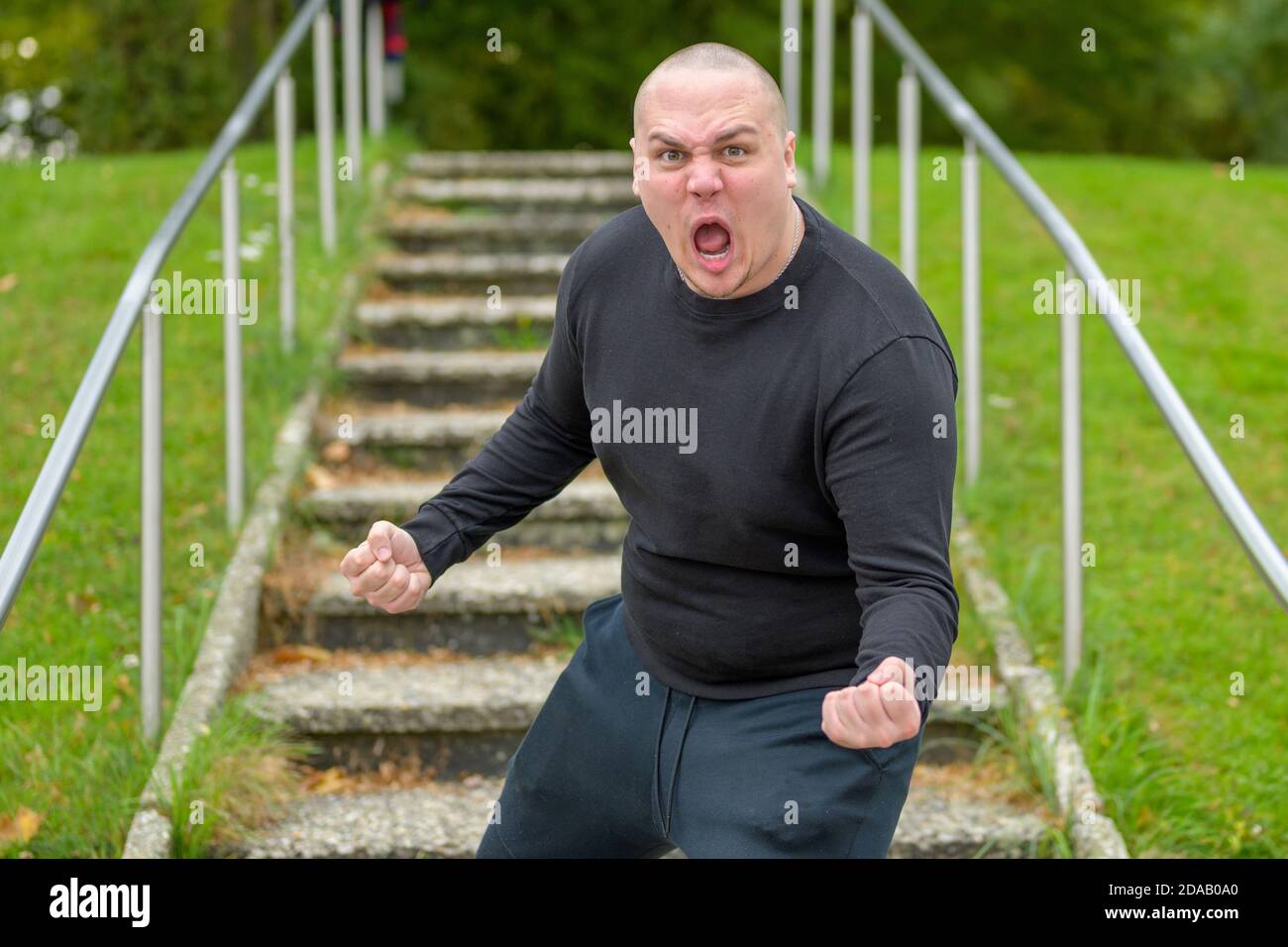 Furious young man yelling at the camera as he stands blocking an exterior set of steps in a park brandishing his fists in anger Stock Photo