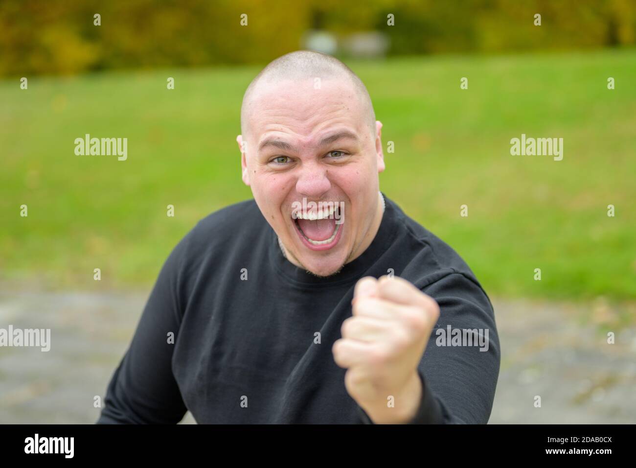 Aggressive threatening angry young man yelling at the camera as he punches with his fist outdoors in a park Stock Photo