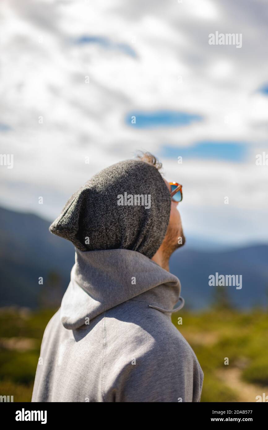 Young white man from behind with sunglasses in beautiful natural setting surrounded by mountains looking up at cloudy sky Stock Photo