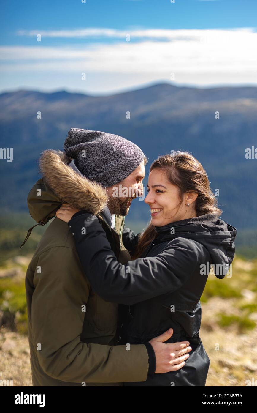 Young couple in love and in winter clothes hug and smile in front of the mountain range in a sunny day Stock Photo