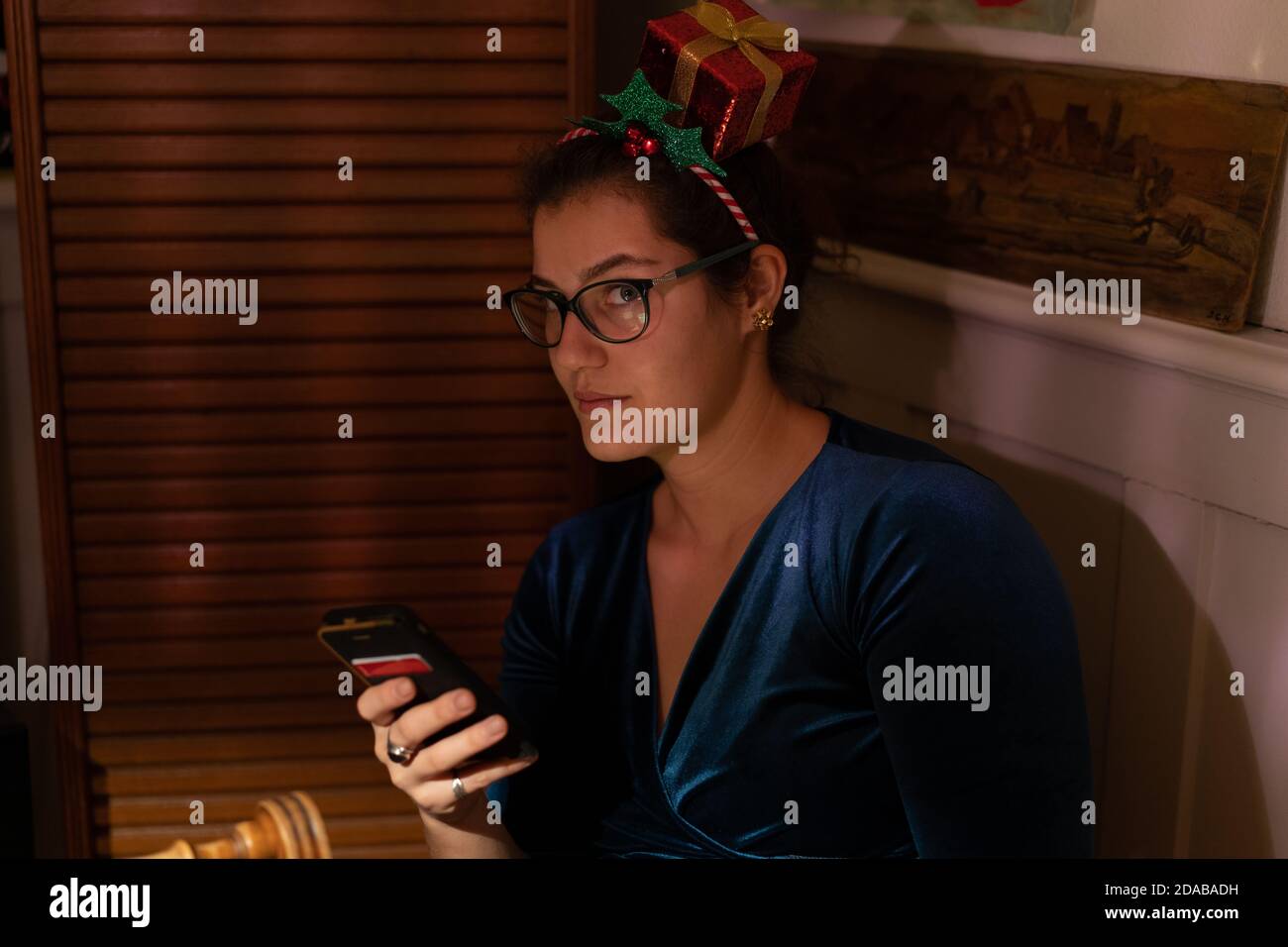 Young woman with christmas hair decor is looking at camera out of the corner of her eyes, holding cellphone at christmas. Stock Photo