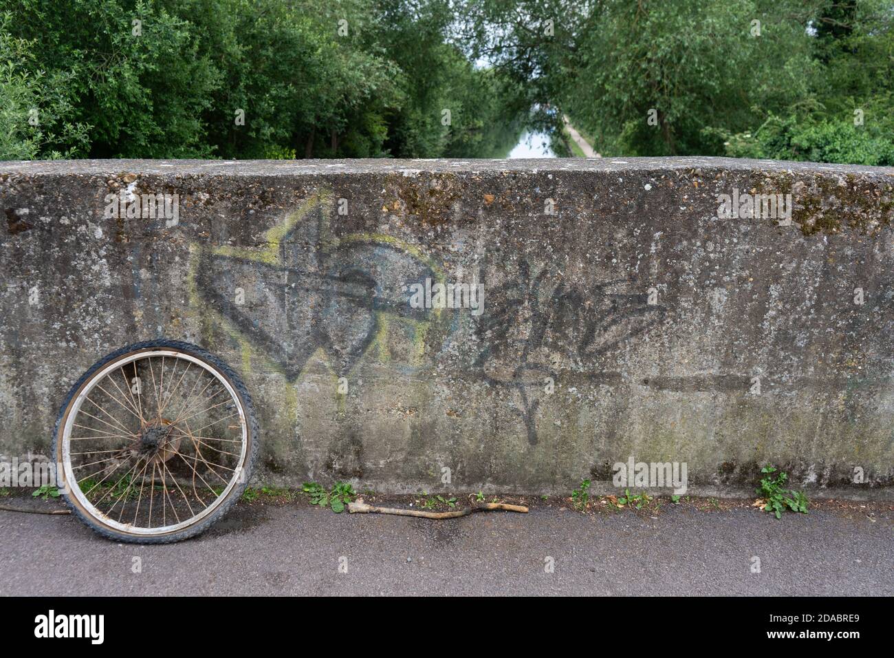 Old rusty bike wheel abandoned on concrete graffiti bridge across canal river lined with beautiful trees Stock Photo