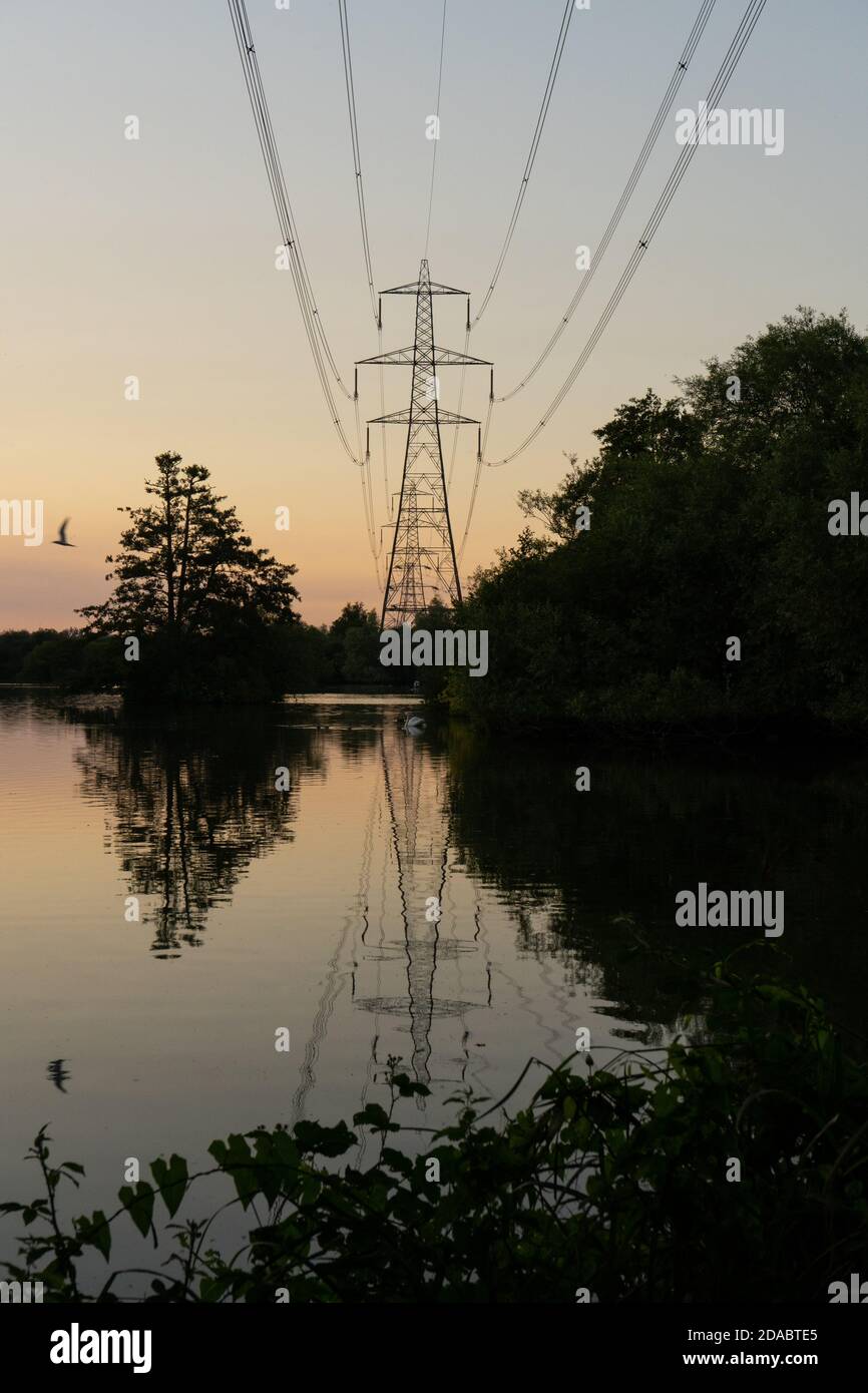 Silhouetted pylon and one flying bird reflected onto beautiful still peaceful lake, at dusk in summertime England. An orange hint of colour, last glim Stock Photo