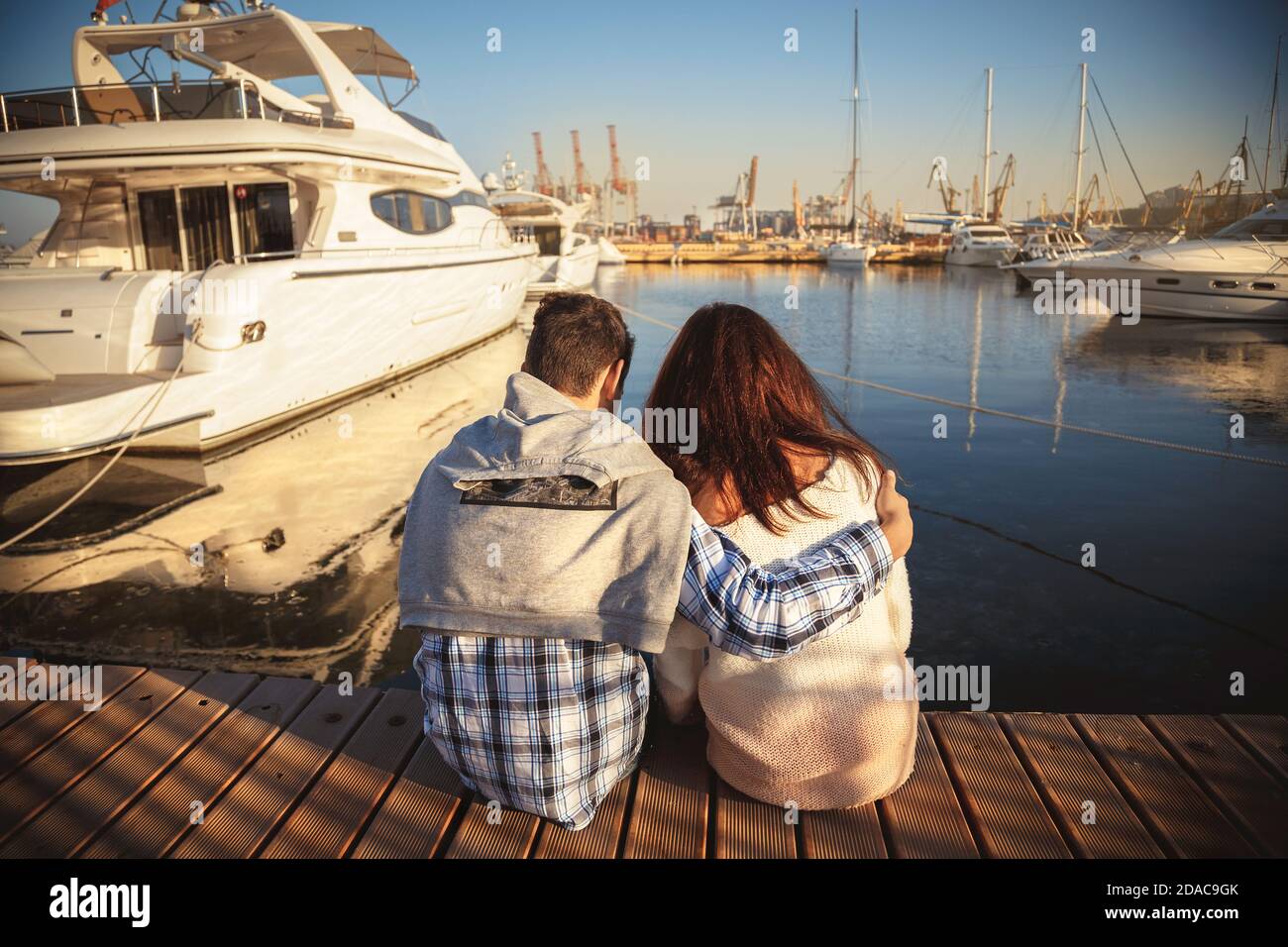 Back view of young couple sitting together on wooden pier in the port with small yachts near to the sea water. Stock Photo