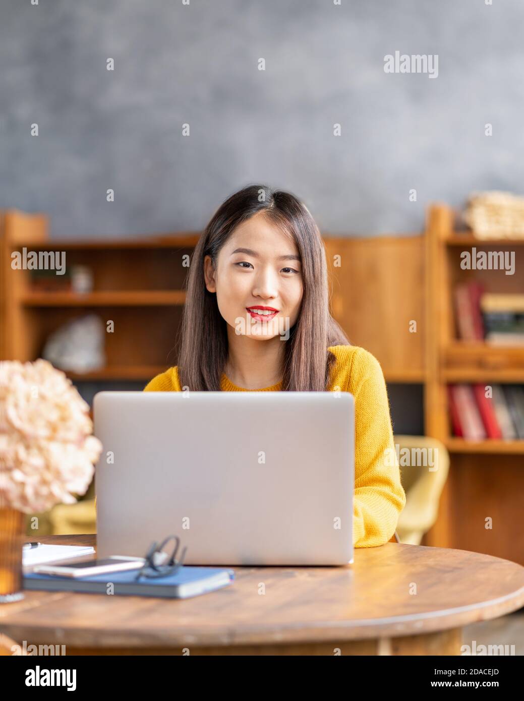 Happy smiling asian woman working on laptop at home or in cafe. Young lady in bright yellow jumper Stock Photo