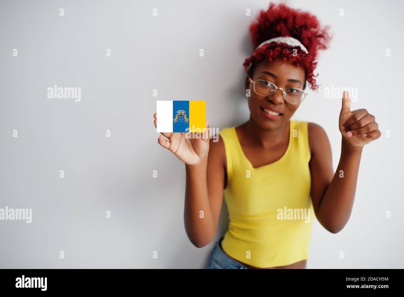 African woman with afro hair, wear yellow singlet and eyeglasses, hold Canary Islands flag isolated on white background, show thumb up. Stock Photo
