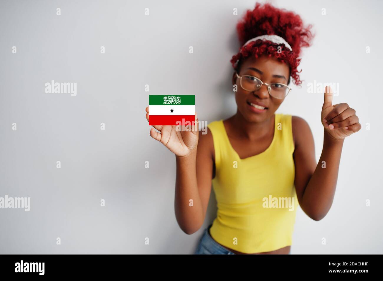 African woman with afro hair, wear yellow singlet and eyeglasses, hold Somaliland flag isolated on white background, show thumb up. Stock Photo