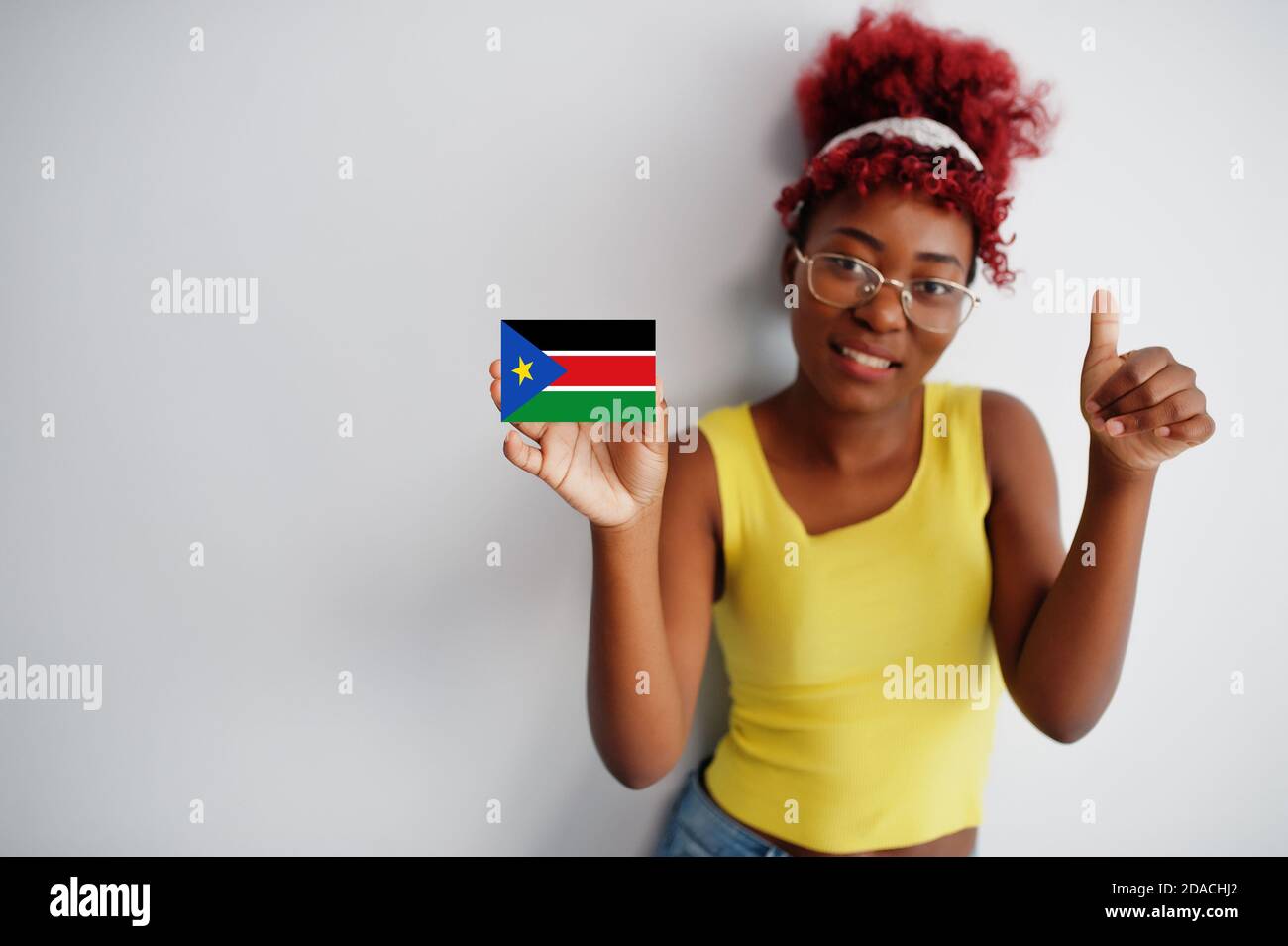 African woman with afro hair, wear yellow singlet and eyeglasses, hold South Sudan flag isolated on white background, show thumb up. Stock Photo