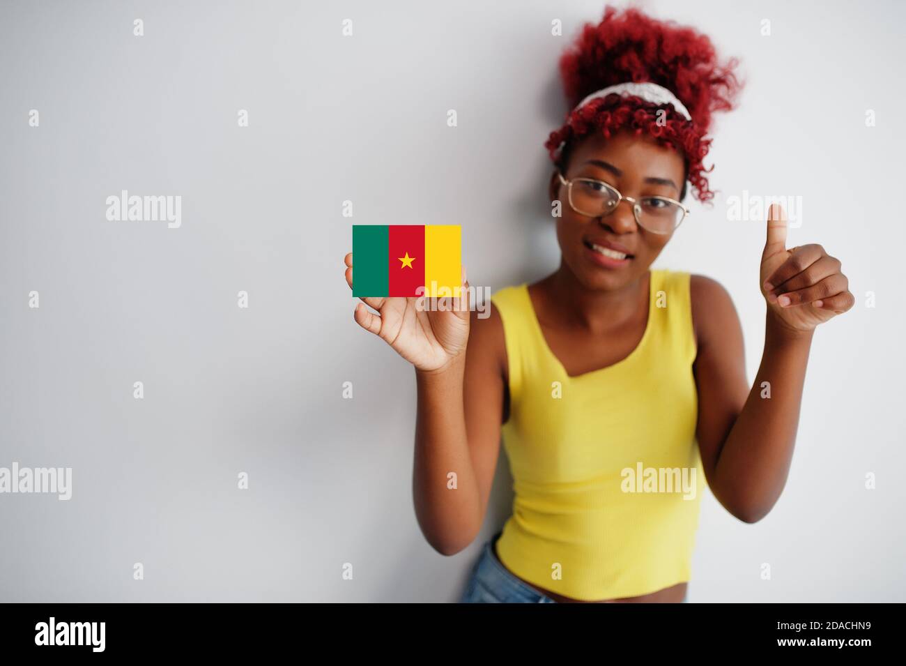 African woman with afro hair, wear yellow singlet and eyeglasses, hold Cameroon flag isolated on white background, show thumb up. Stock Photo