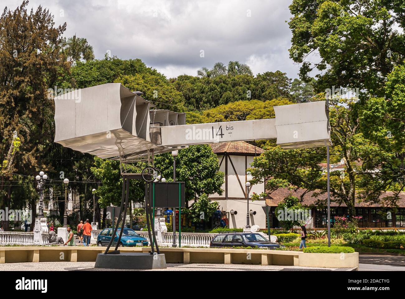 Petropolis, Brazil - December 23, 2008: Gray statue of flimsy historic flying machine in park full of green trees. Cars and people around, under gray Stock Photo