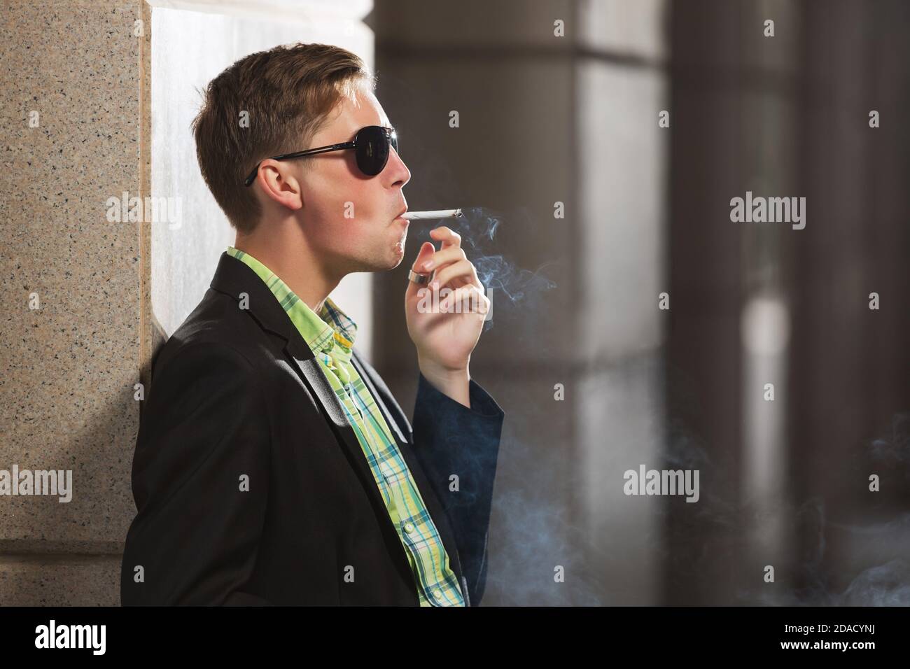 Young man in black blazer and sunglasses smoking a cigarette on city street Stock Photo
