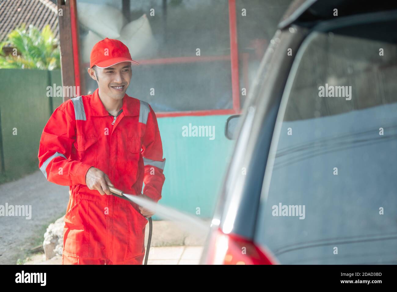 smiling male car cleaner wearing a red uniform and a hat while spraying