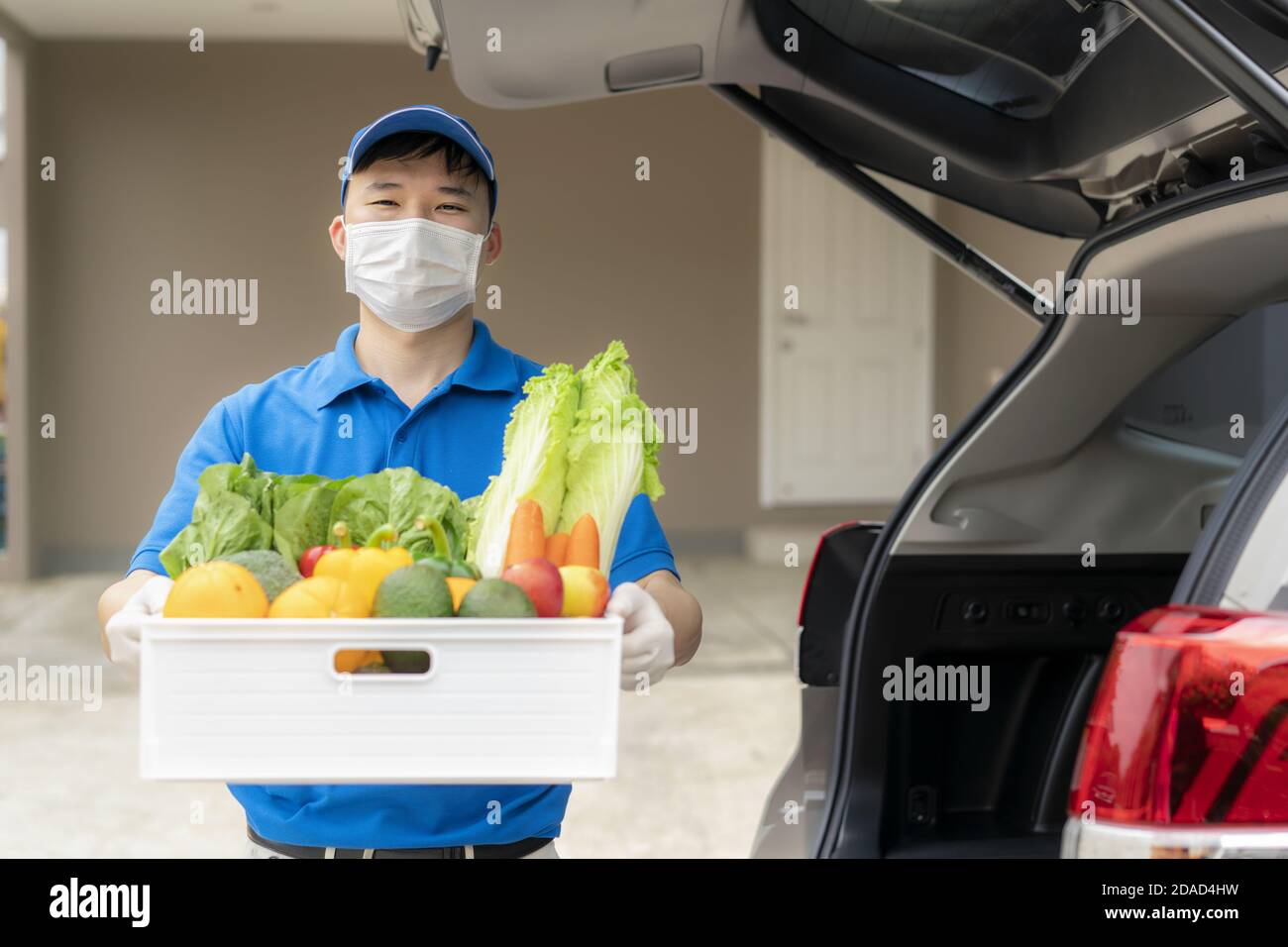 Asian delivery man wearing face mask and glove with groceries box of food, fruit, vegetable and drink standing near van in front of the customer house Stock Photo