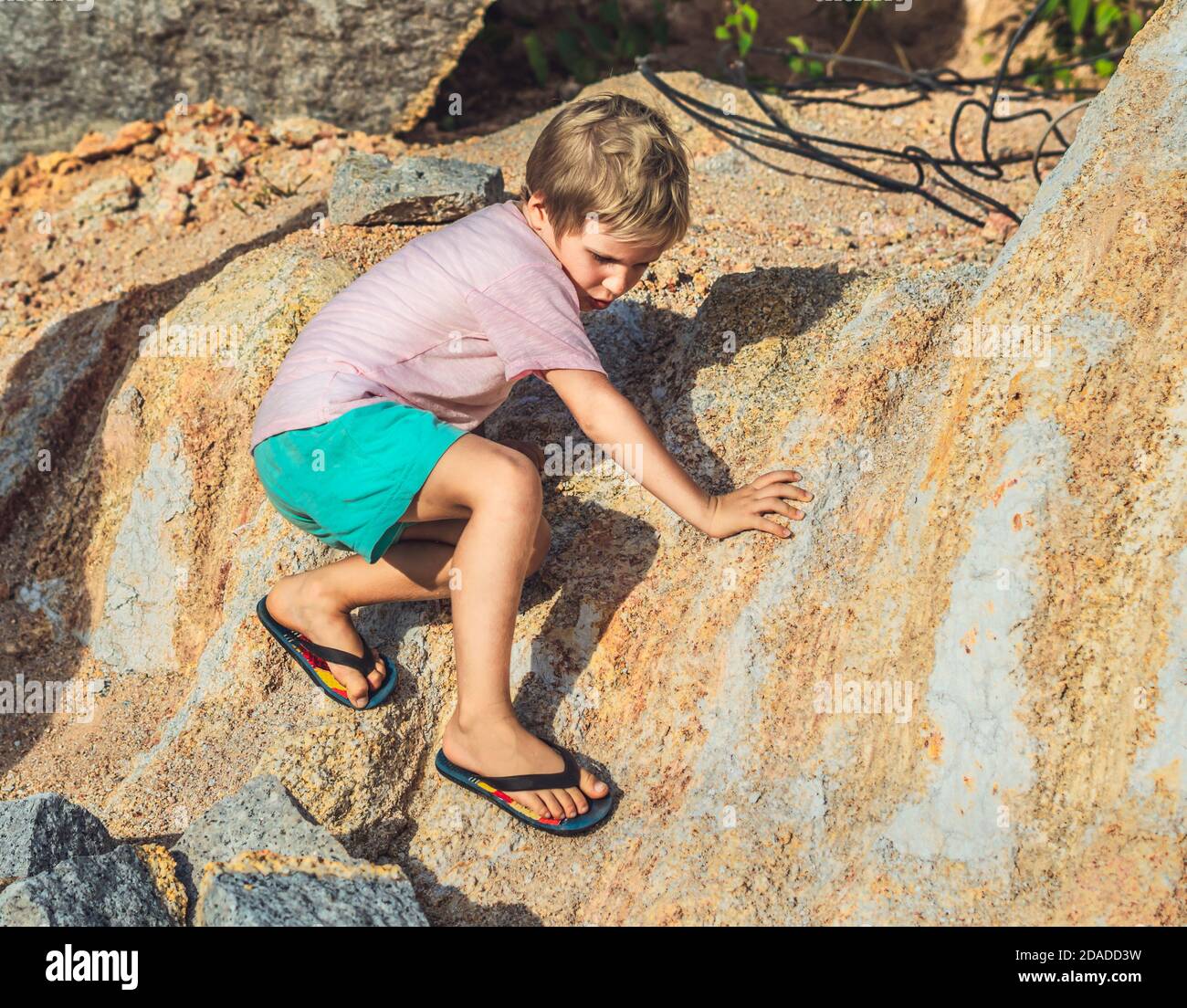 Mischievous hooligan playful boy climbs rock stone in flip flops. Choose foot wear for nature sport game. Warn children about safety danger outdoor Stock Photo