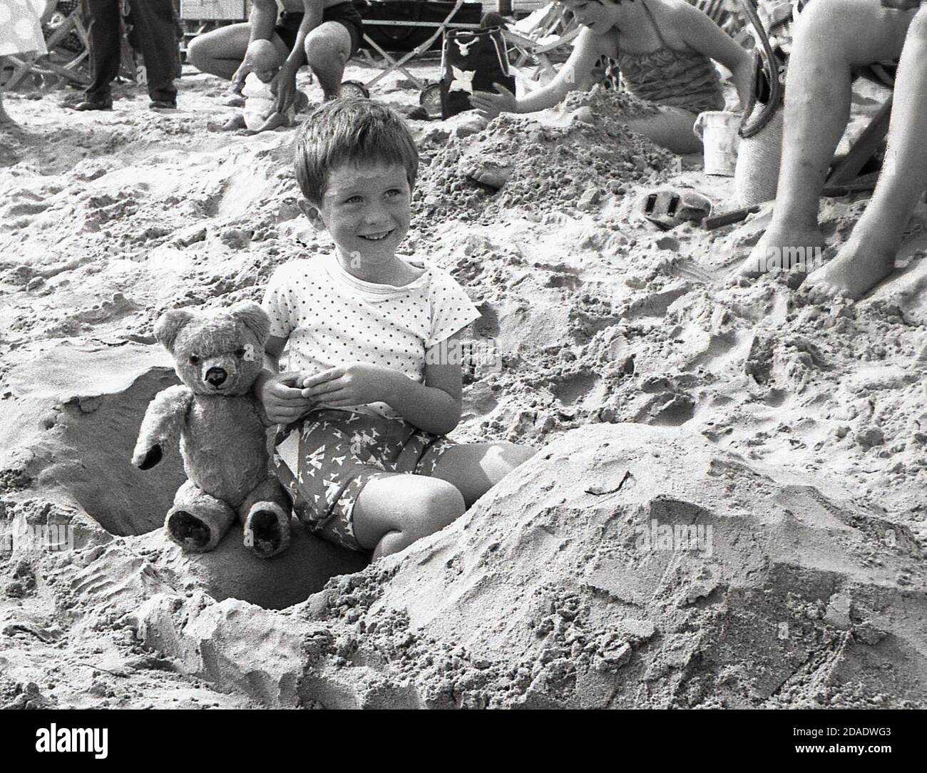 1960s, historical, sweet little boy sitting on a sandy beach ion his sandcastle with his teddy bear, England, UK. Stock Photo