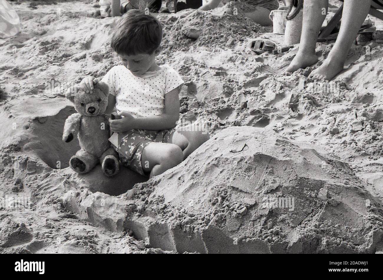 1960s, historical, sweet little boy sitting on a sandy beach ion his sandcastle with his teddy bear, England, UK. Stock Photo