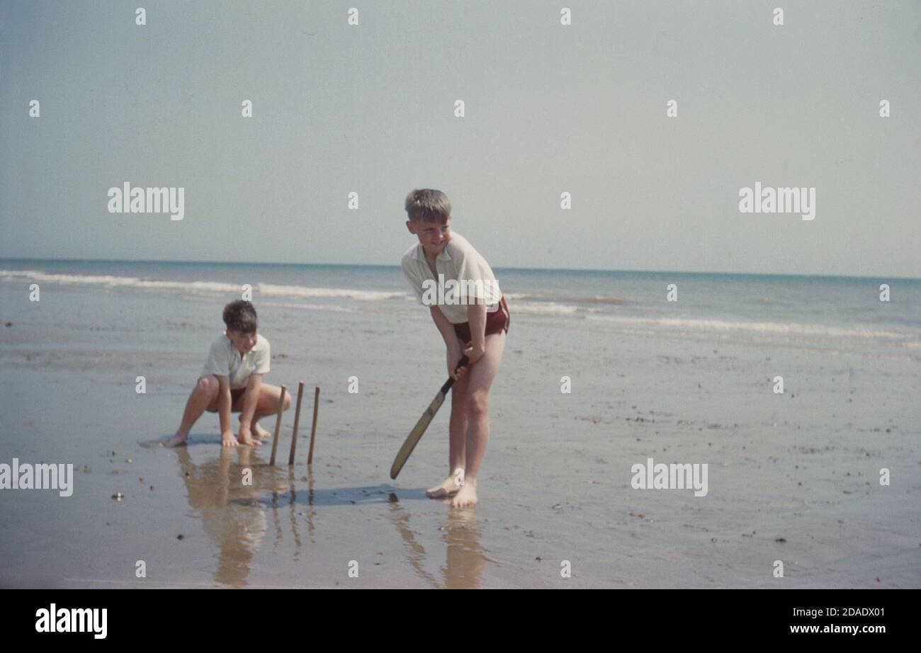 1960s, historical, two young boys in white tops and swimming trunks, playing a game of beach cricket outside on a flat, wet sandy area by the sea, England, UK. One is batting, the other is behind the make-shift stumps, wicket-keeping. In beach cricket the batter is obliged to run whenever the ball is hit regardless of how good a contact was made. A hit into the sea is generally regarded as six and out! Stock Photo