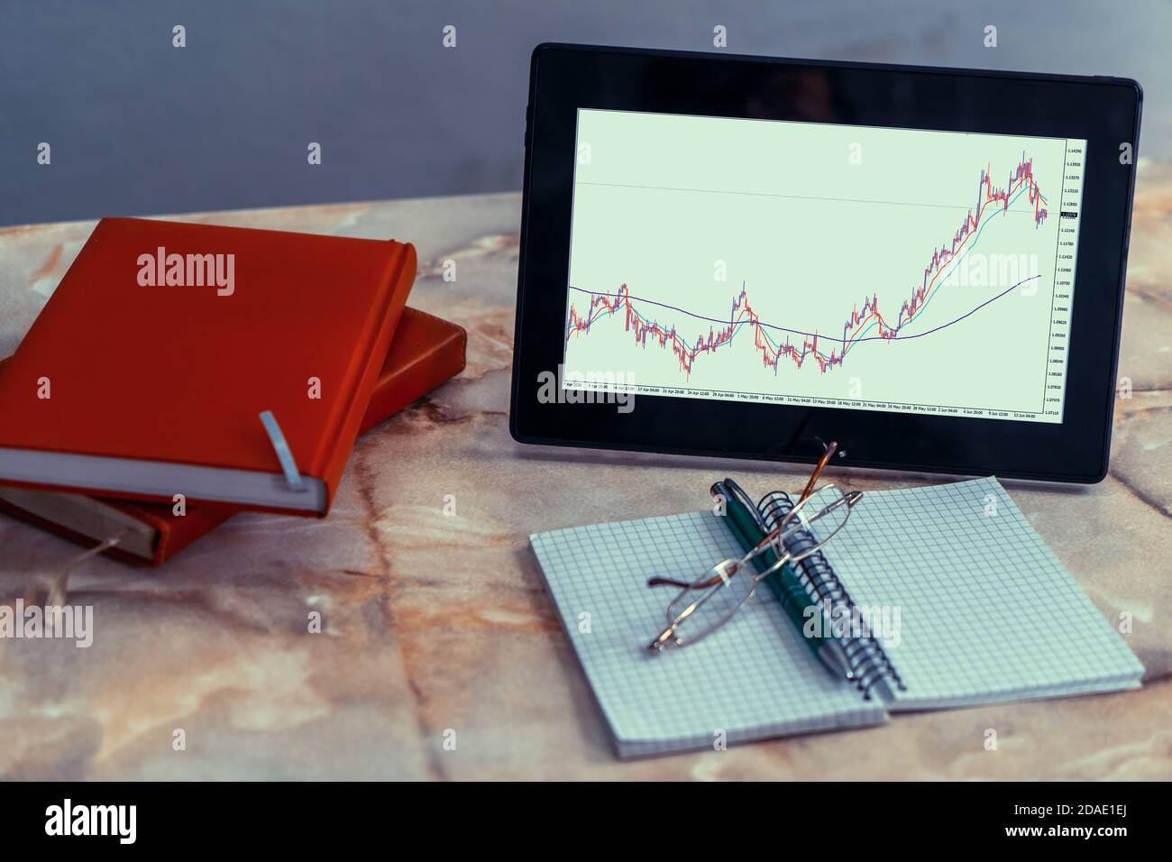 A tablet screen on a desk displaying a currency pairs chart. There are a notebooks, a pen and a glasses too. Stock Photo