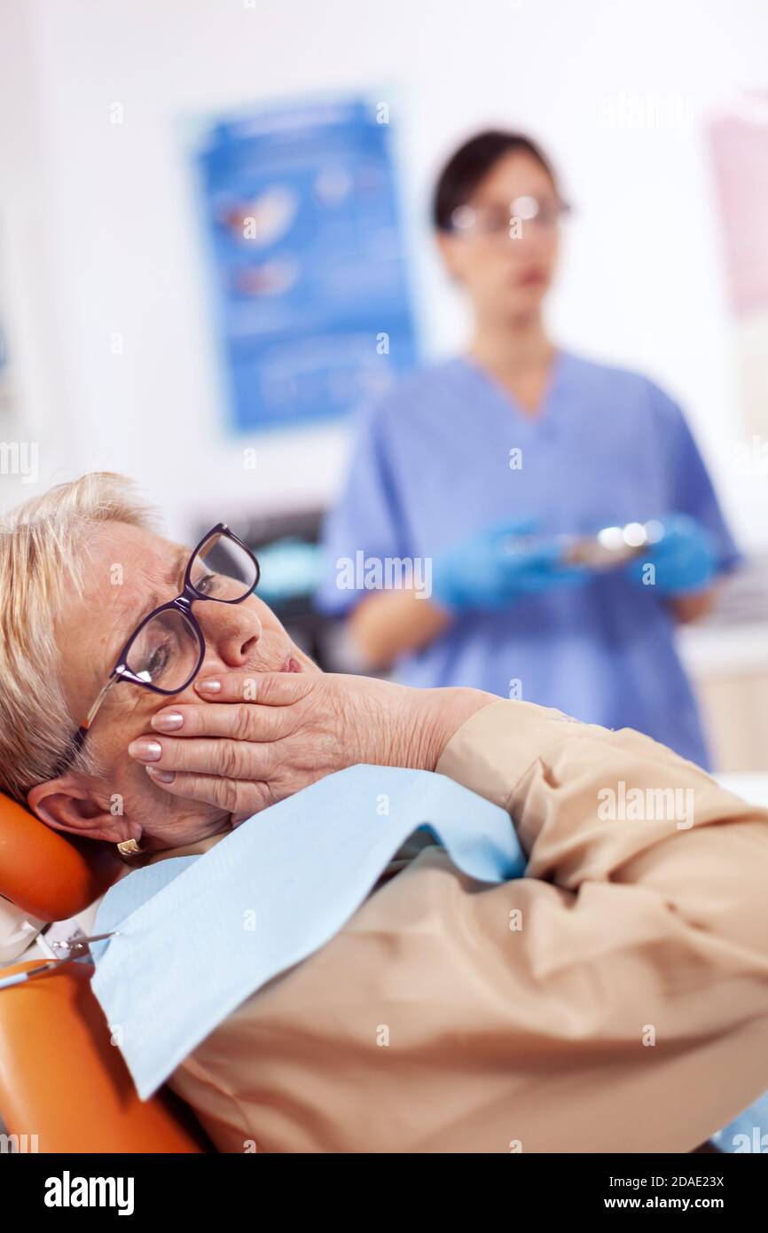 Middle age patient touching mouth with painful expression sitting on chair in dentist cabinet. Senior woman in healthcare hospital accusing and complaining about tooth. Stock Photo