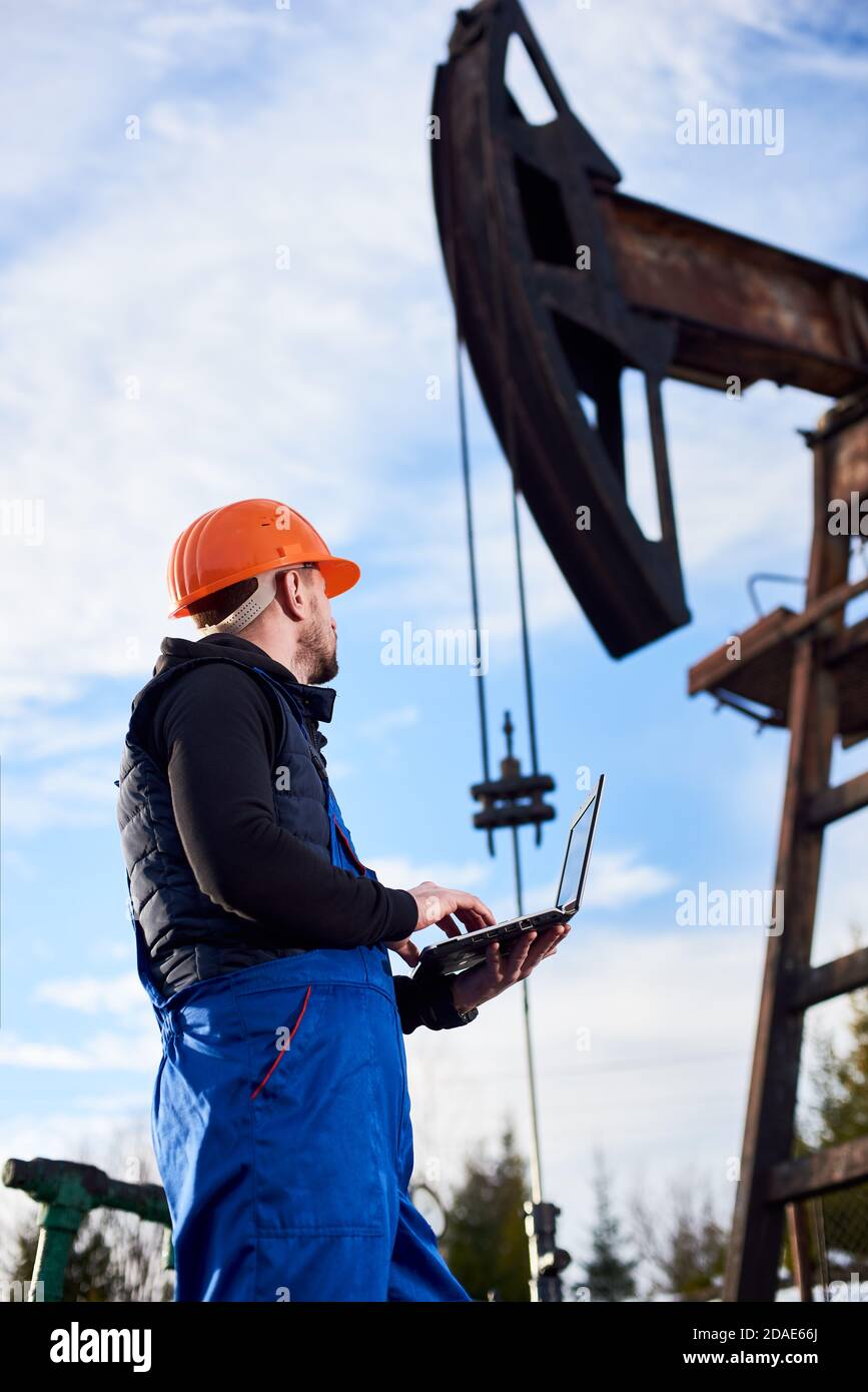 Oil man holding notebook and looking at oil pump rocker-machine. Male worker in uniform and helmet using laptop and controlling work of pump jack. Concept of petroleum industry and oil extraction. Stock Photo