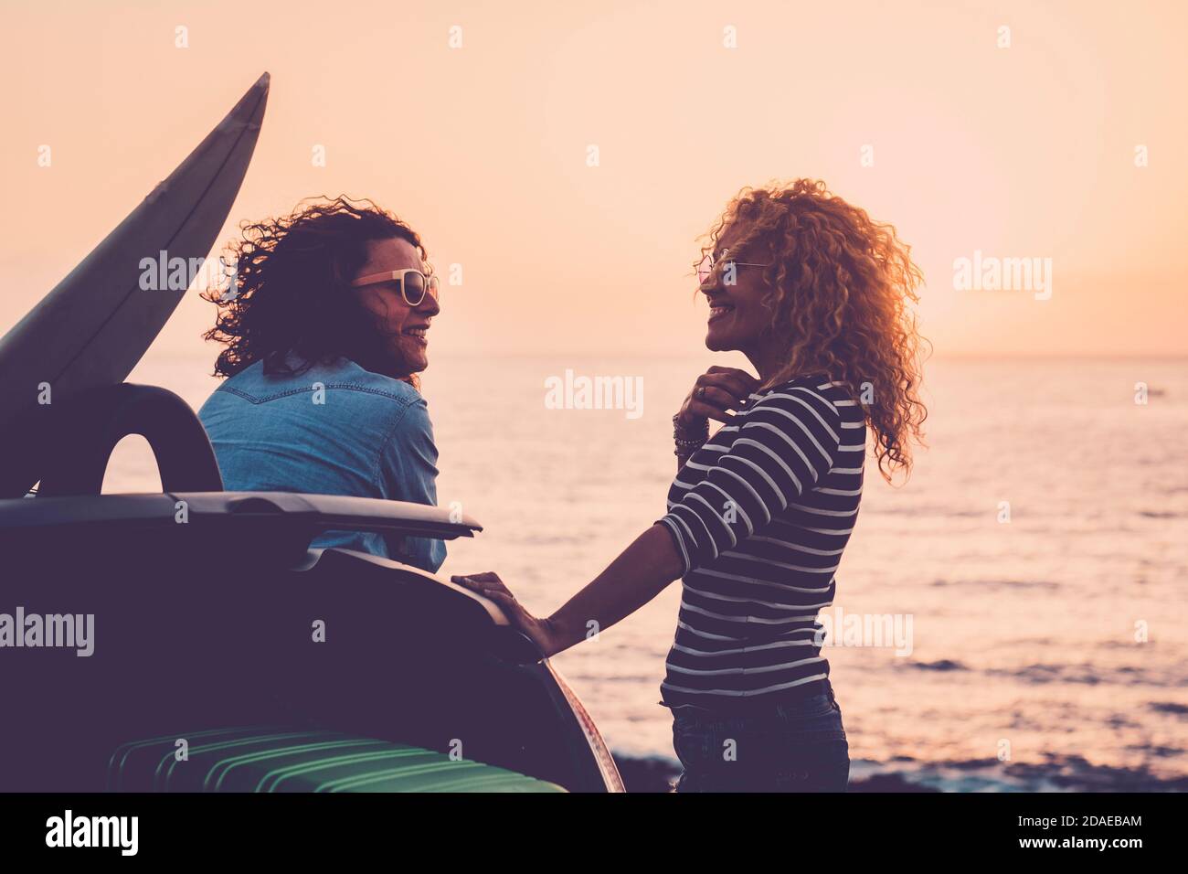 Cheerful and happy couple of women friends enjoying the sunset and travel together for vacation time - sea in background and young middle age enjoyed people near a car and surf table - concept of friendship and travel Stock Photo