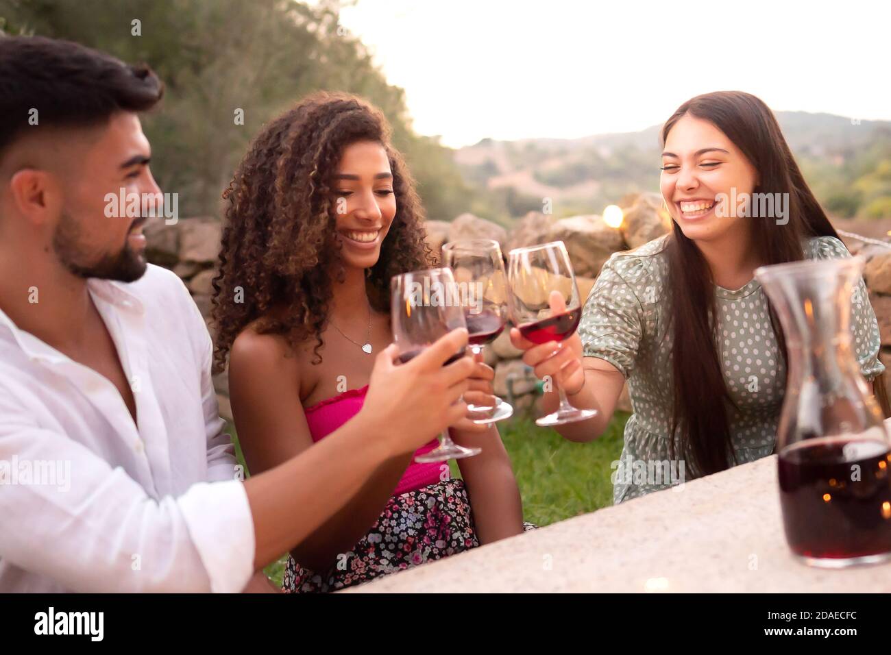 Three friends clinking glasses outdoor at sunset - Mixed race person group toasting with red wine glasses in countryside for the vineyard harvest end Stock Photo