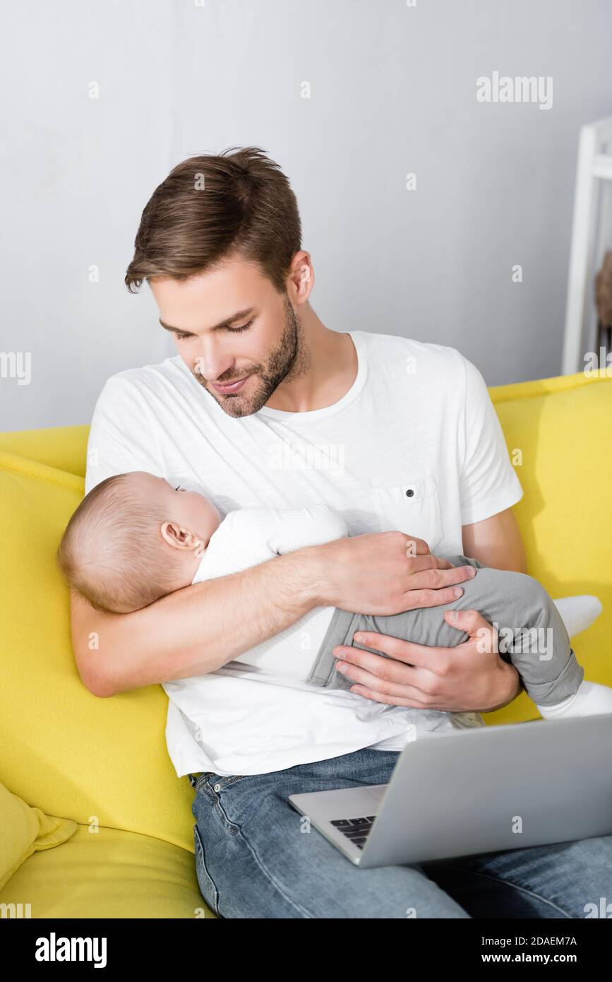 caring father holding in arms sleepy baby son while sitting on sofa with laptop Stock Photo