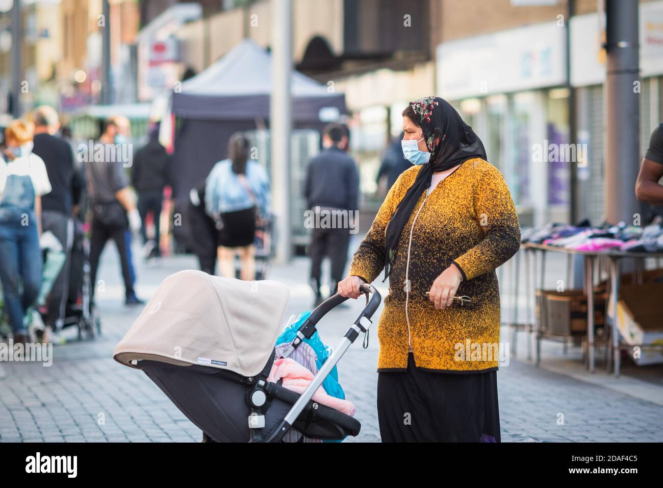London, UK - 3 November, 2020 - A woman wearing a face mask and headscarf with a baby stroll while shopping at Walthamstow market Stock Photo