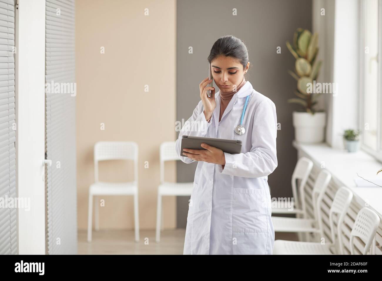 Young female doctor in white coat talking on mobile phone and using digital tablet for online work standing at the corrior at hospital Stock Photo