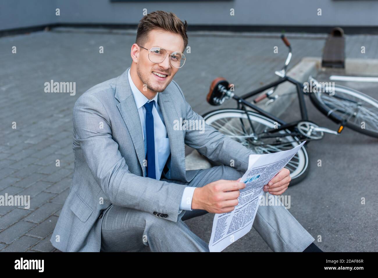 Smiling businessman holding newspaper near bicycle on blurred background outdoors Stock Photo
