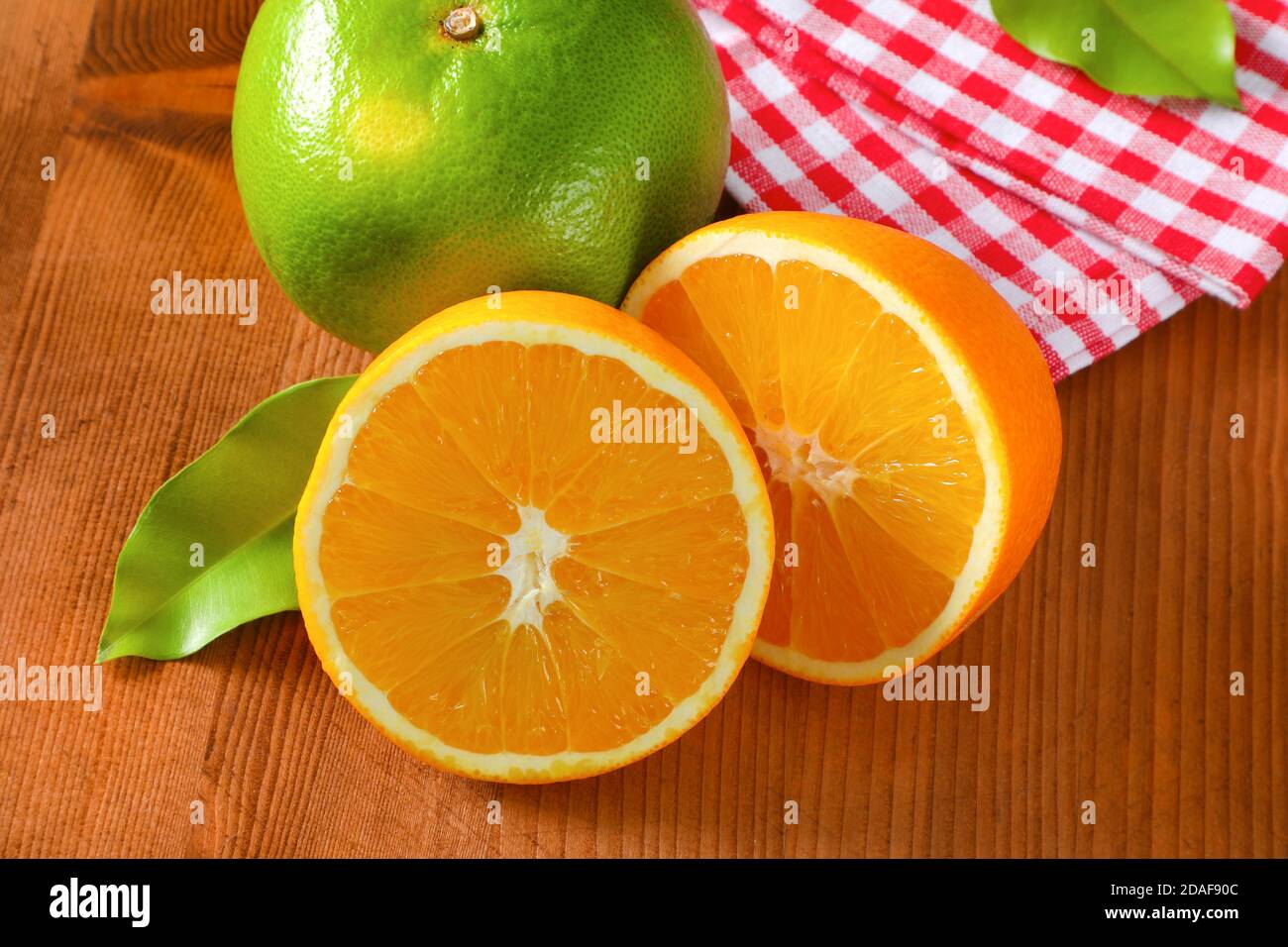 Green grapefruit (sweetie, pomelit, oroblanco), two orange halves and checked red and white tea towel on wooden table Stock Photo