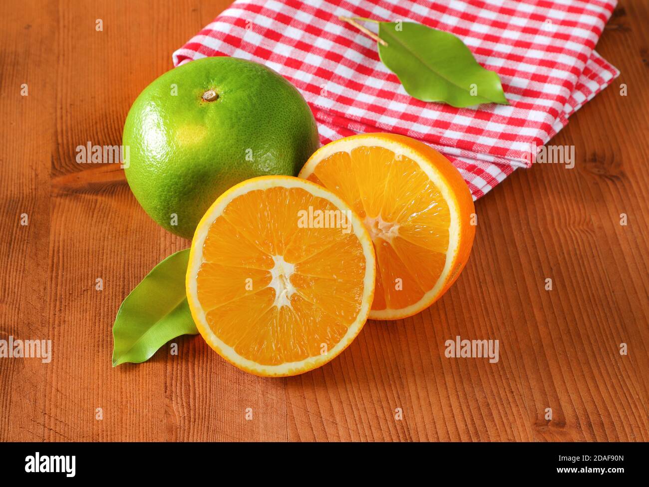 Green grapefruit (sweetie, pomelit, oroblanco), two orange halves and checked red and white tea towel on wooden table Stock Photo