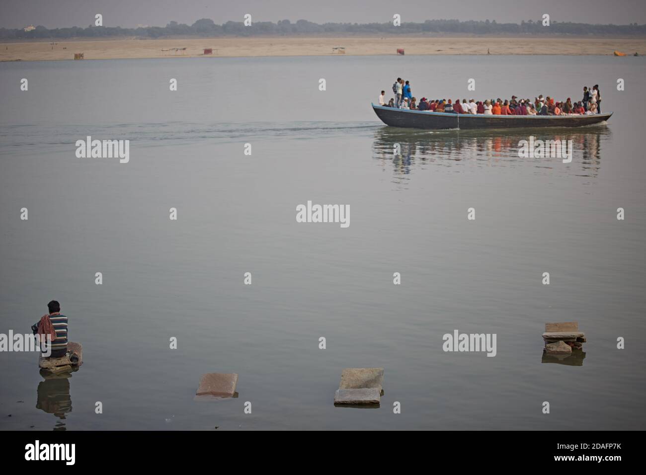 Varanasi, India, December 2015. A boat full of pilgrims passes in front of a man sitting on a stone to wash clothes in the Ganges River. Stock Photo