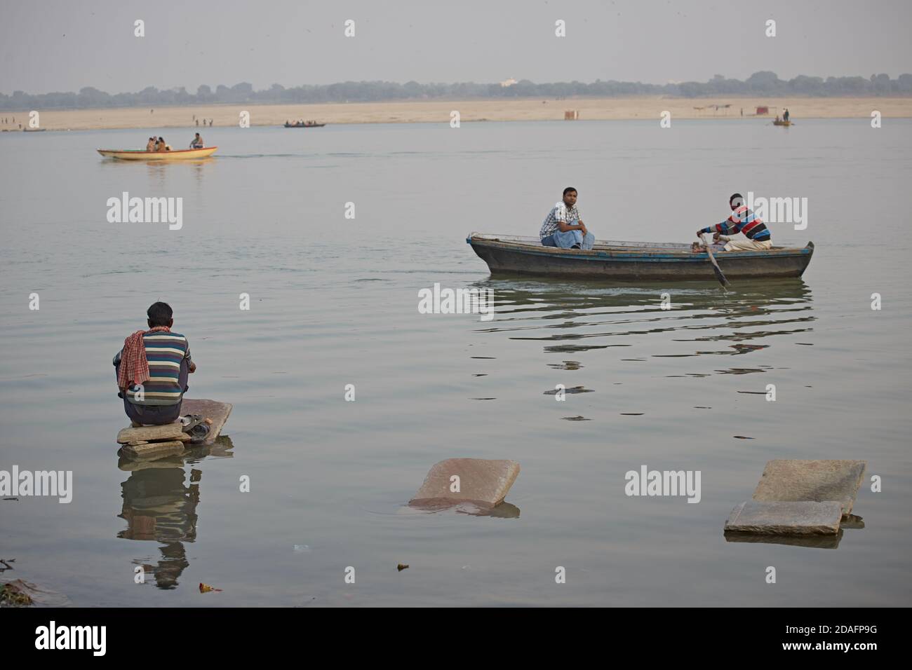 Varanasi, India, December 2015. A rowboat passes in front of a man sitting on a washing stone in a Ganges river ghat. Stock Photo