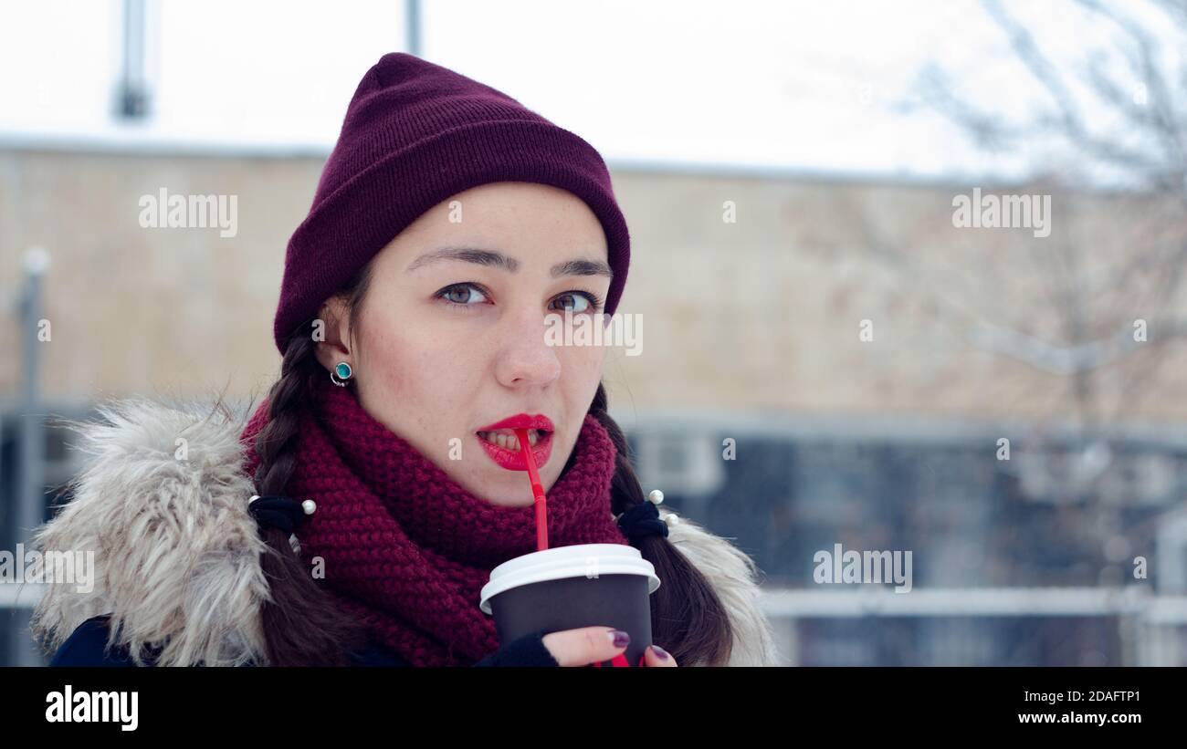 Beautiful young girl in a romantic mood with pleasure drinks coffee through a straw in a winter park. Stock Photo