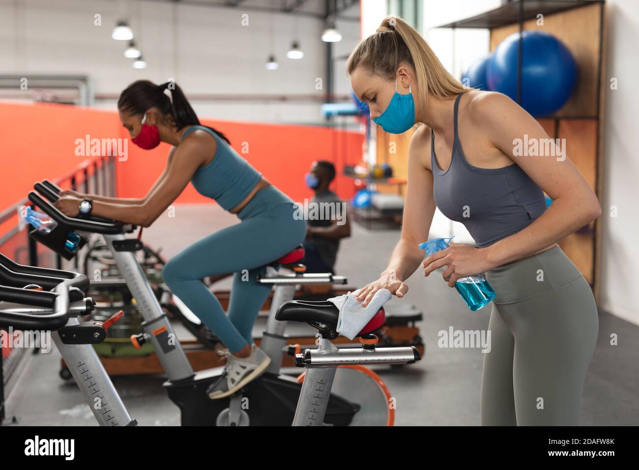 Fit caucasian woman wearing face mask sanitizing seat of stationary bike before working out in the g Stock Photo