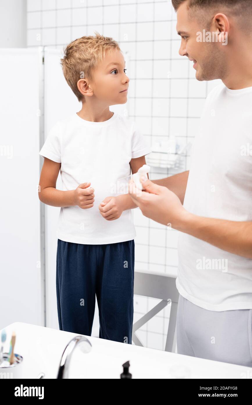 Excited preschooler boy looking at father with dental floss holder while standing on chair in bathroom Stock Photo