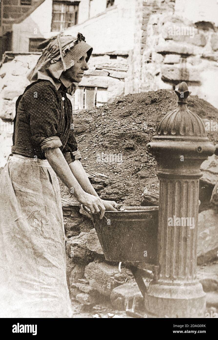 A Victorian photograph of a Woman collecting water at the Cragg Pump, Whitby, North Yorkshire in the late 1800s. Steps led down from the pump onto Pier road where fishwives would 'gip' or gut herrings as they arrived on the quay directly from the fishing boats. The pump was then the only source of fresh water in the area. The headress she is wearing is known as a 'Steerths' Bonnet, Taking its name from the village of Staithes where they were manufactured, though they were widely worn amongst the fishing communities elsewhere. Stock Photo