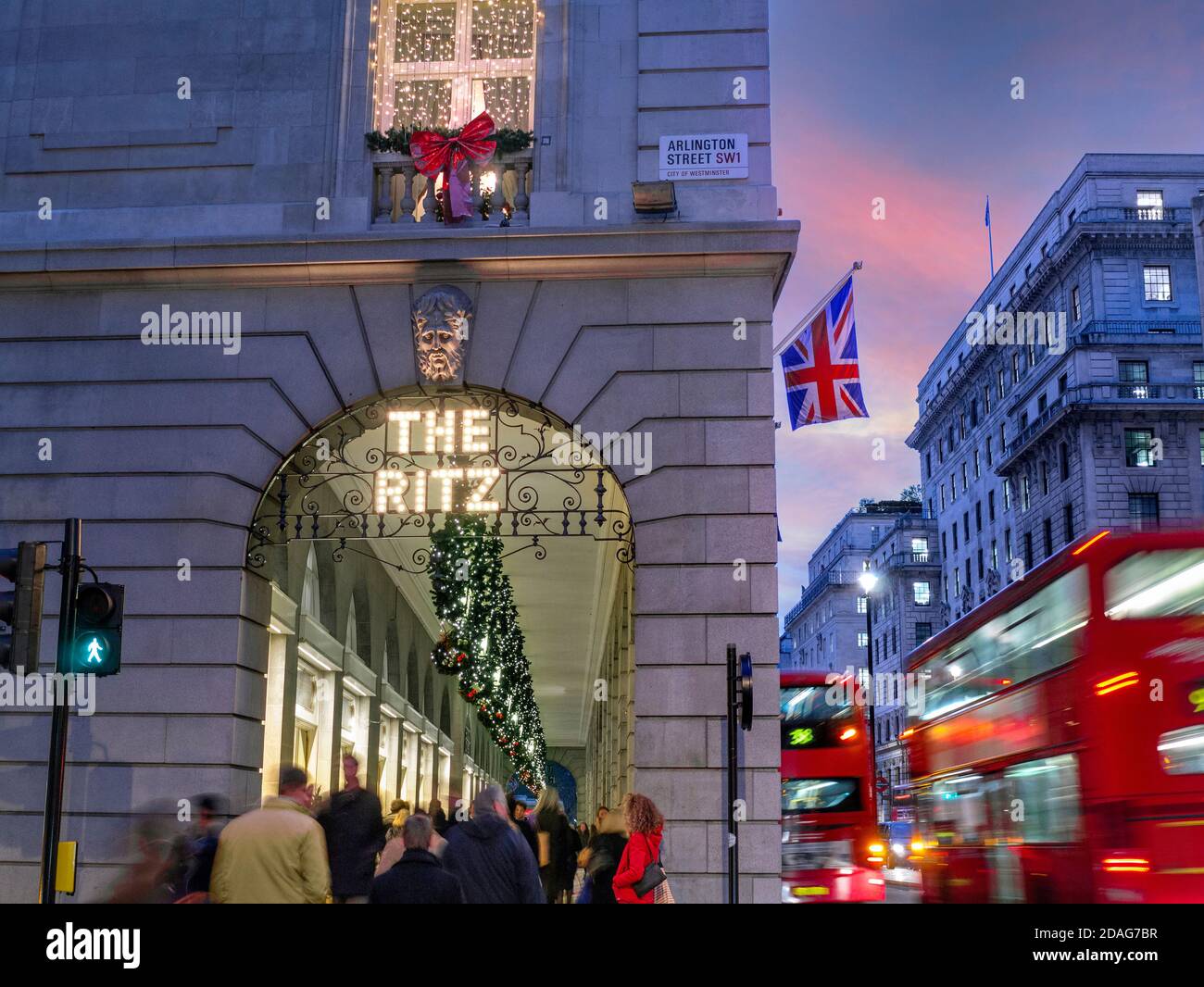 LONDON CHRISTMAS CROWDS The Ritz Hotel at winter busy festive season, evening night lights ‘The Ritz’ sign illuminated, with a Union Jack flag, shoppers and passing blurred London red buses Arlington Street Piccadilly London UK Stock Photo
