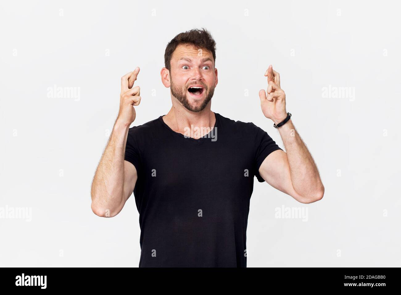 Crossed fingers. Amazing and cheerful smiling man in black t-shirt studio shot, isolated on white Stock Photo