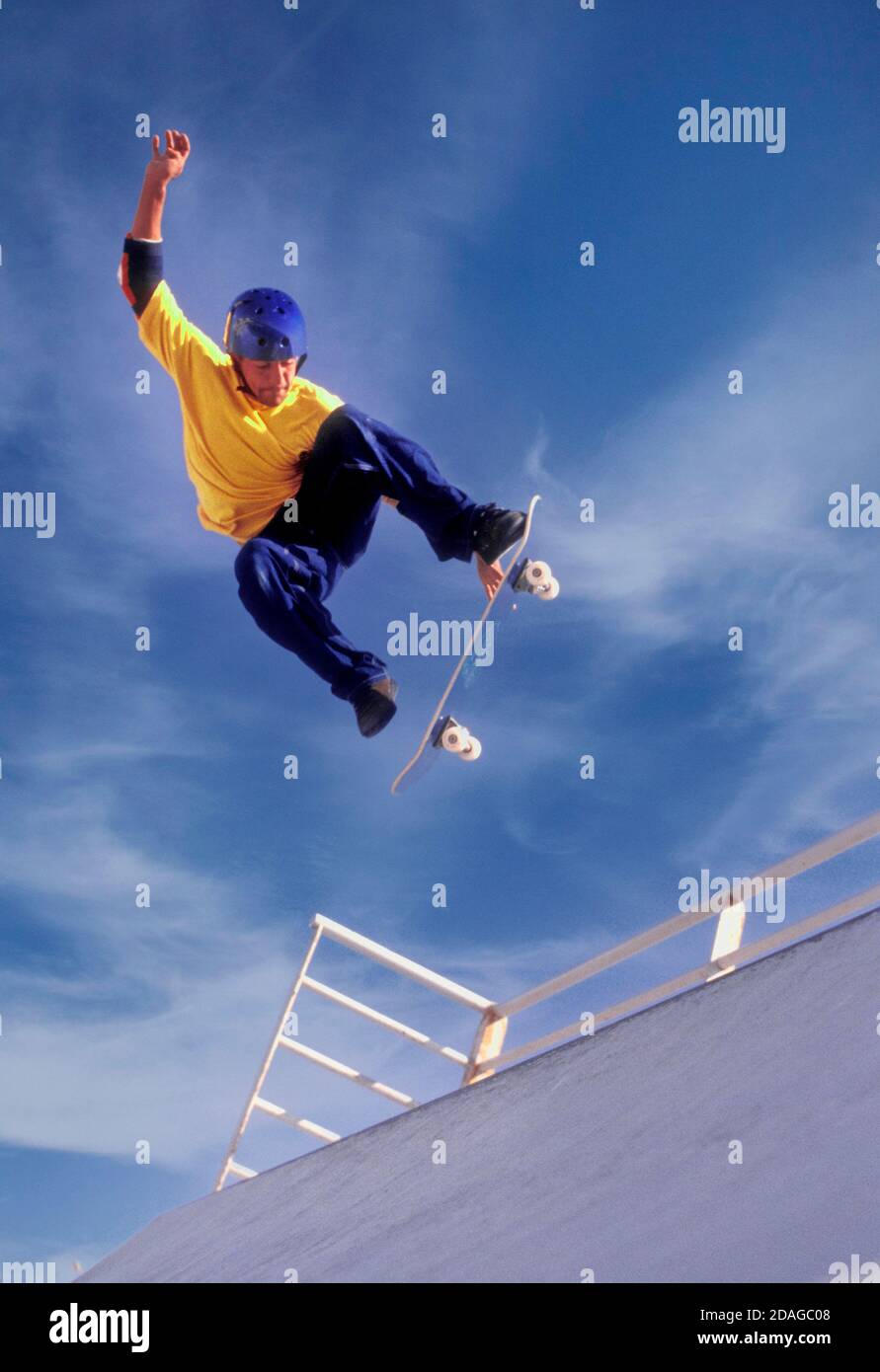 Skateboarder wearing helmet and protective armbands, captured in midair flying against a sunny blue sky Stock Photo