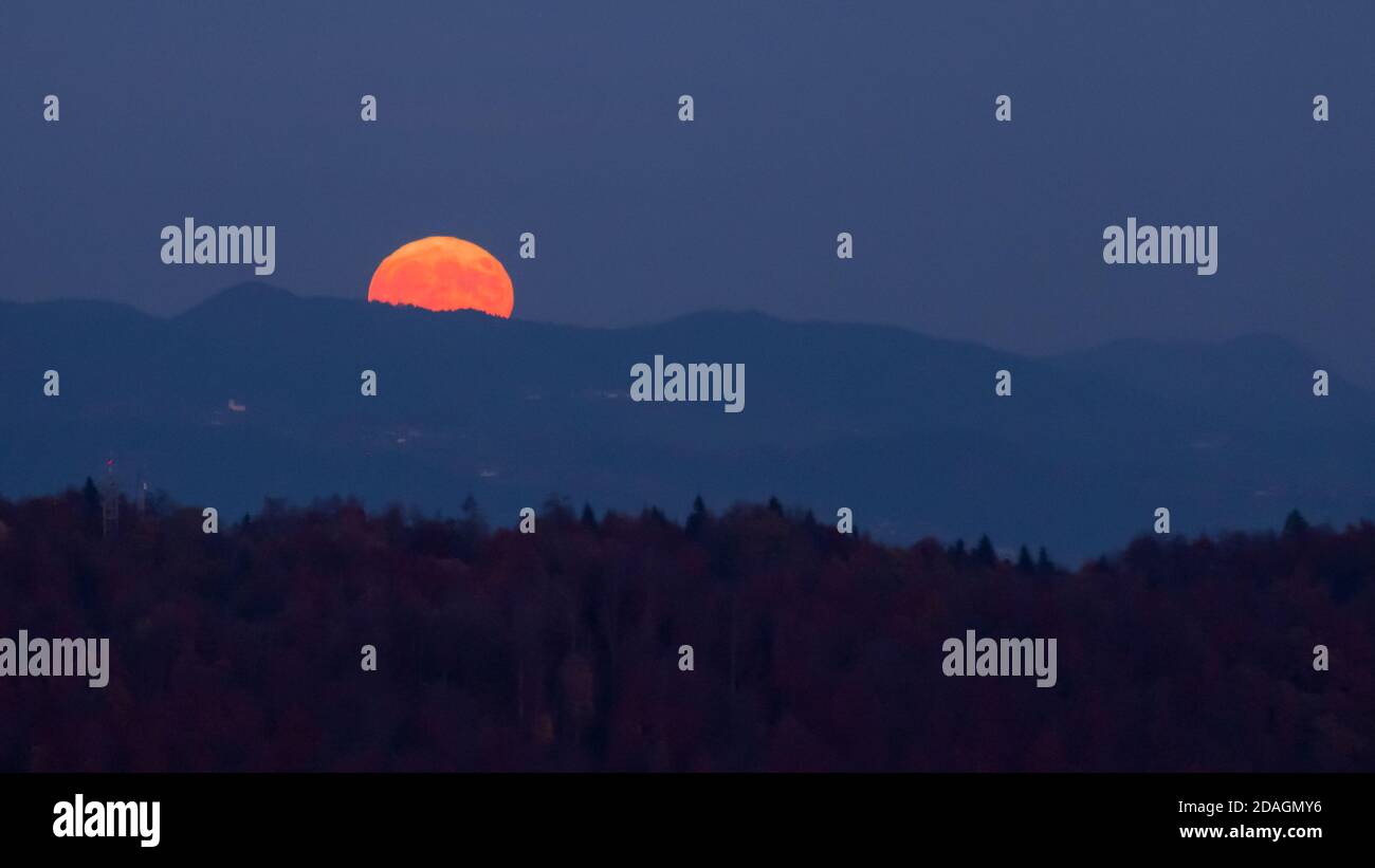 huge orange full moon rising into the night sky behind a mountain ridge with layers of hills in the foreground creating a mystical atmosphere Stock Photo