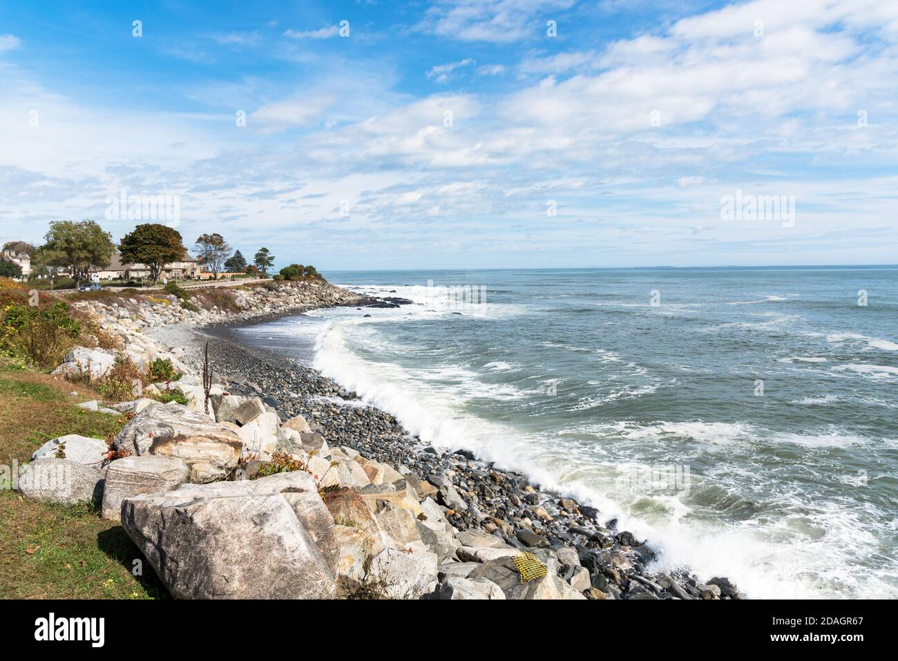 Small pebble beach along the coast of New Hampshire on a sunny autumn day. A clifftop coastal road lined with houses is visible in background. Stock Photo