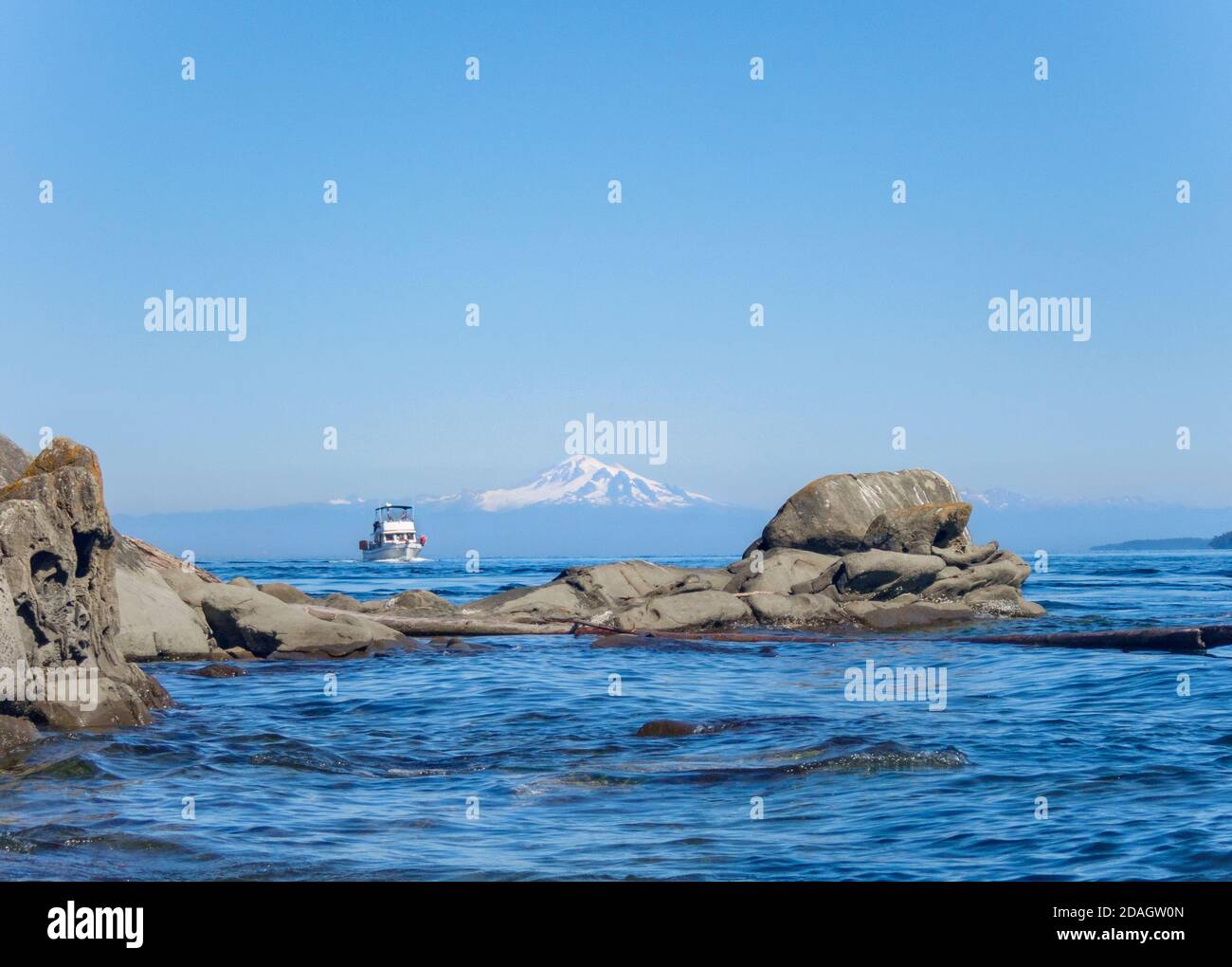 An eroded sandstone reef frames a view of a motor yacht approaching from the Strait of Georgia and in the distance, Mt. Baker's snowy peak Stock Photo