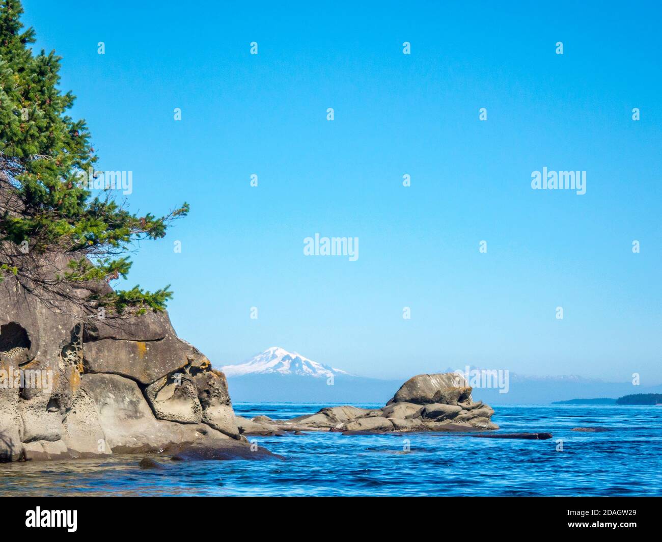 A reef extends out from an eroded island shoreline in the Strait of Georgia, framing a view of Mt. Baker, whose snowy peak rises in the distance. Stock Photo