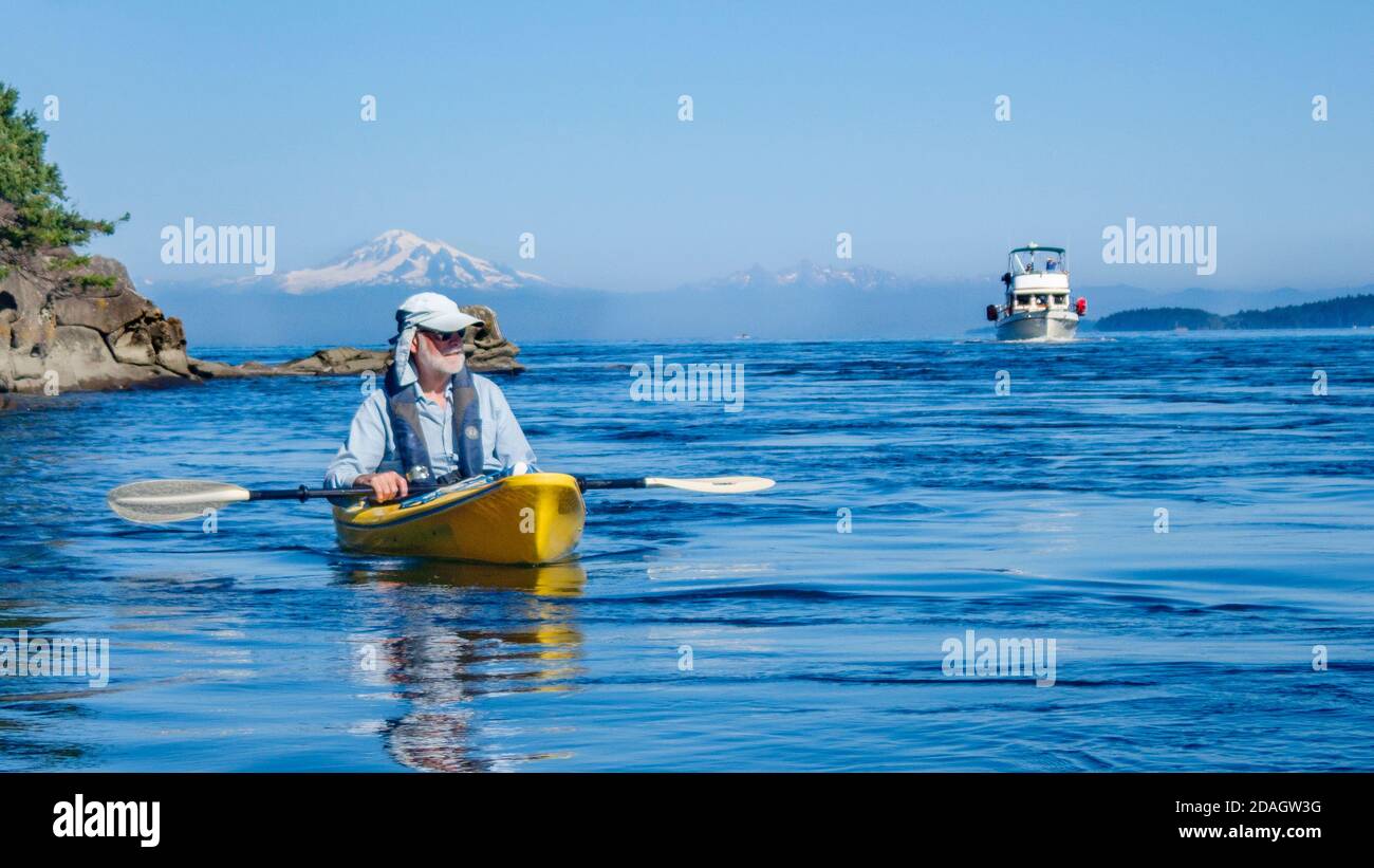 On a summer day in the Salish Sea, a man in a sun hat watches from his kayak as a motor yacht approaches, with Mt. Baker's snowy peak in the distance. Stock Photo