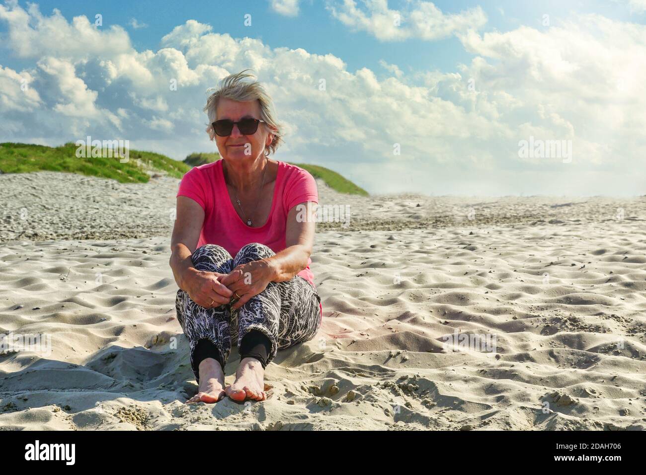 Elderly caucasian woman sitting on beach sand with hands around knees. Full length. Stock Photo