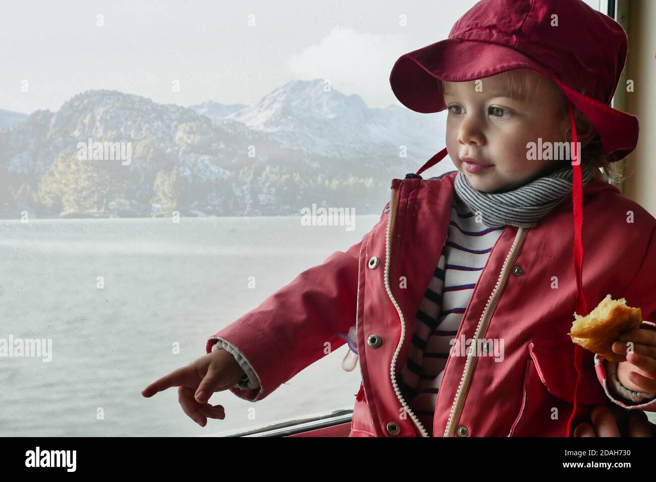 Toddler dressed in coat and hat optioning and looking away. Lake and mountain scenic background. Medium shot. Stock Photo